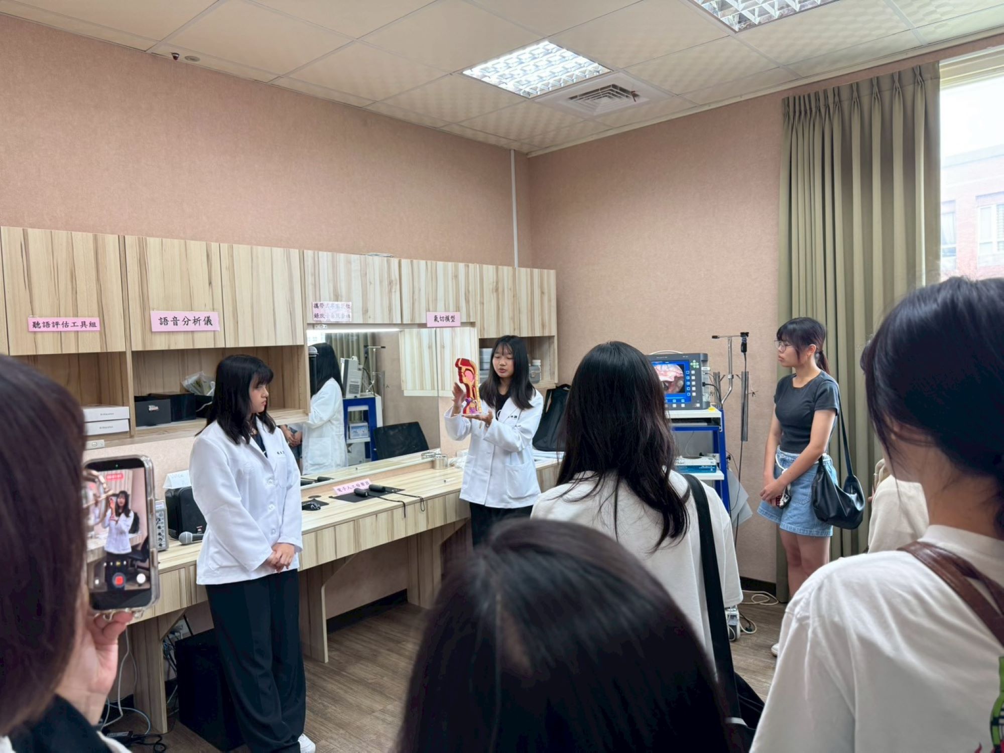 在嗓音吞嚥實驗室中,學姊手持精細的解剖喉部模型,解碼語言治療的核心專業,考生也以手機記錄專業場景。