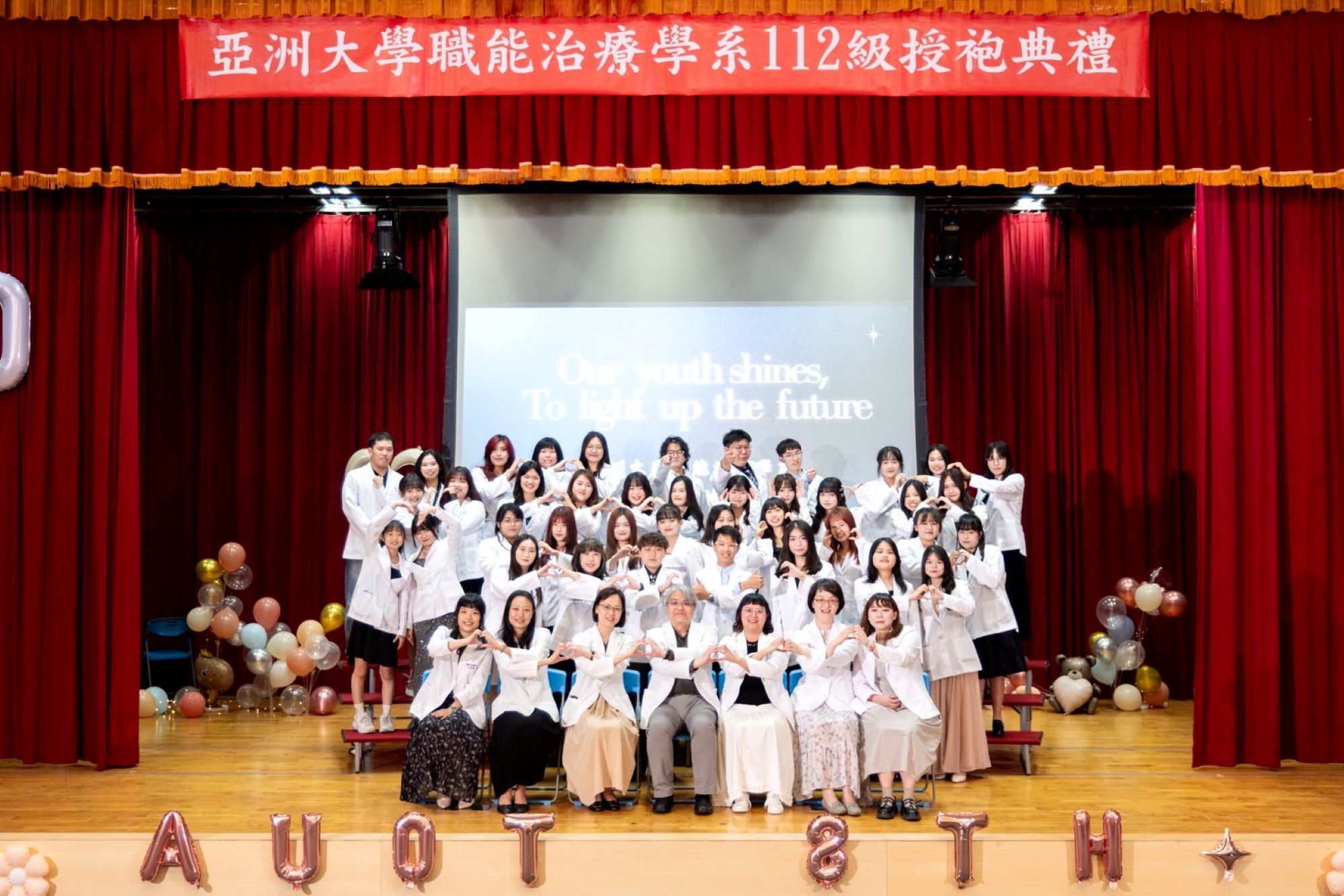 亞大職治系授袍典禮舞台大合照,師生身穿白袍溫馨比心,共慶專業傳承。