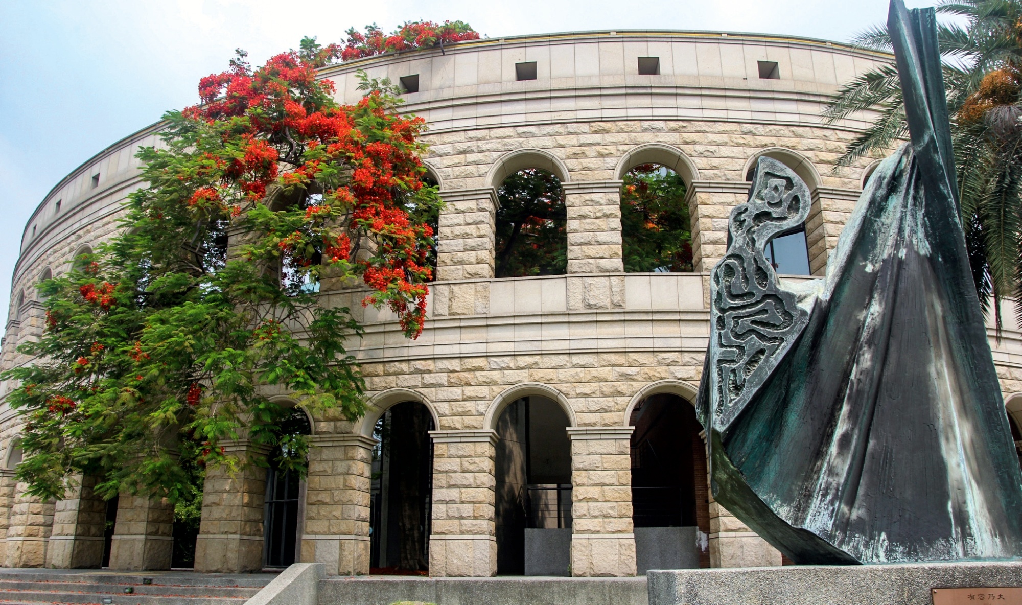 亞大體育館羅馬式建築旁，火紅鳳凰花熱情盛開，與青銅雕塑交織出初夏迷人的校園風光。